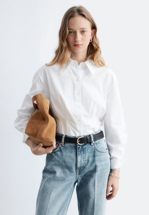 Mujer joven vestida con una camisa blanca de botones y jeans azul claro, sosteniendo un bolso de gamuza color beige bajo el brazo, sobre un fondo liso.