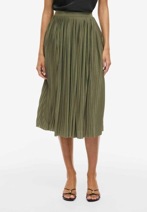 Kvinne iført en olivengrønn plissért midi-skjørt kombinert med svarte sandaler med tynne stropper og åpne tær.