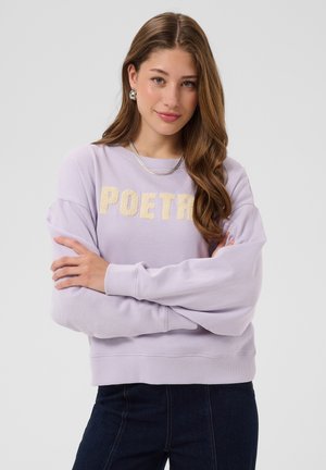 Kvinde med langt brunt hår bærer en lilla sweatshirt med teksten ""POETRY"", mørke jeans, et sølvhalskæde og store øreringe, armene er krydsede.