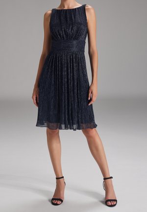 Femme portant une robe sans manches bleu marine plissée mi-longue avec une large ceinture et des talons hauts noirs avec brides à la cheville, debout sur fond gris.