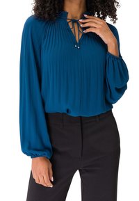 Blouse teal à manches longues en tissu plissé. Dotée d'un col en V avec un détail de nœud et de manches ballon. Portée avec un pantalon noir.