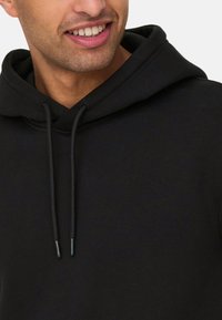 Hoodie noir avec cordon de serrage, fabriqué en tissu doux. Comprend une poche avant et des poignets côtelés. Design minimaliste, sans motifs visibles.