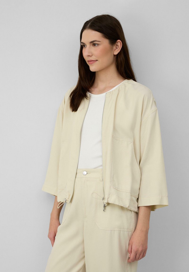 Beige, ritsjack met een ontspannen pasvorm en brede driekwartmouwen. Voorzien van trekkoorden aan de onderkant en voorzakken.