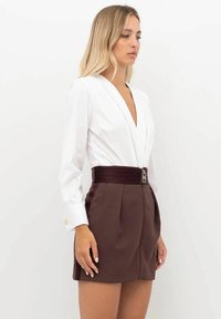 Donna bionda che indossa una camicetta bianca a maniche lunghe infilata in una gonna marrone scuro a vita alta con cerniera frontale e ampia cintura.