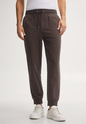 Mann trägt dunkelbraune gemusterte Jogginghose mit Kordelzug in der Taille und weiße Sneakers, steht auf hellem Hintergrund.