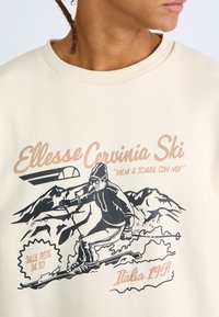 Sudadera beige con un gráfico de esquí, ilustración de contorno negro y texto retro, con cuello redondo acanalado y textura suave.