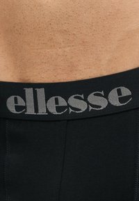 Muški donji dio leđa vidljiv je iznad crnih bokserica sa sivim logom „ellesse“ na pojasu.