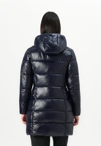 Vrouw met haar rug naar de camera, gekleed in een lange, glanzende marineblauwe pufferjack met een capuchon, tegen een effen witte achtergrond.