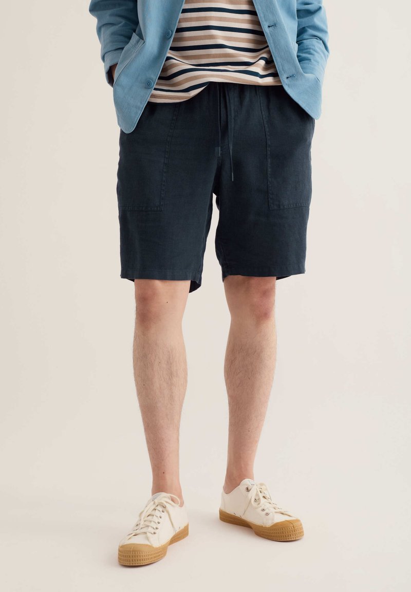 Jambes d'une personne portant un short bleu marine, des baskets beiges avec semelles en gomme, une chemise rayée et une veste bleu clair, debout les mains dans les poches.