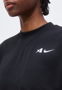 Άτομο που φοράει μαύρο φούτερ με λαιμόκοψη, με λευκά λογότυπα Nike και Air στο αριστερό μέρος του στήθους, ορατό από τον λαιμό έως τον άνω κορμό.