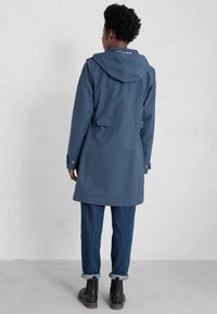 Granatowa wodoodporna parka z kapturem, o dopasowanym kroju, z guzikami i dwoma bocznymi kieszeniami, noszona na zwijanych dżinsowych spodniach i butach.