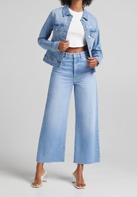 Kvinna iklädd ljusblå denimjacka och vida jeans med råa kanter, vit croppad topp och sandaler med klara klackar, stående mot grå bakgrund.