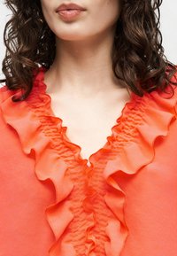 Κοντινό πλάνο γυναίκας με καστανά σγουρά μαλλιά που φοράει πορτοκαλί μπλούζα με βολάν κατά μήκος του V-λαιμού.