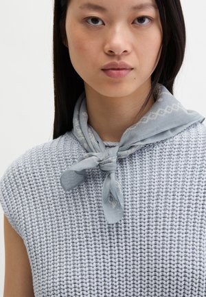 Jonge vrouw draagt een lichtblauw gebreid mouwloos topje en een bijpassende sjaal met patroon om haar hals gebonden, neutrale achtergrond.