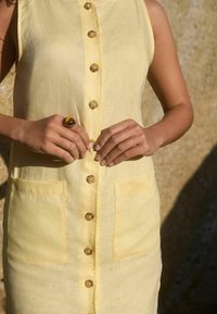 Gelbes Leinenkleid mit Knopfleiste, zwei seitlichen Taschen und glänzenden runden Knöpfen. Glatte Textur, ärmelloses Design.