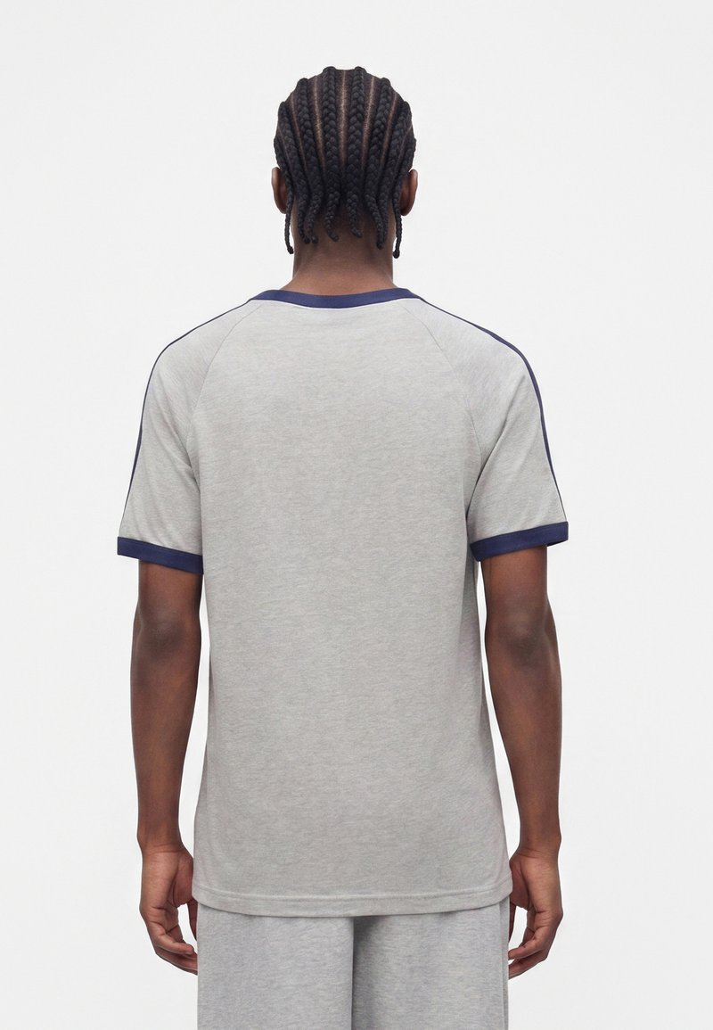 Mann mit Cornrow-Zöpfen, der ein hellgraues Kurzarmshirt mit marineblauem Besatz trägt, steht mit dem Rücken zu einem schlichten weißen Hintergrund.