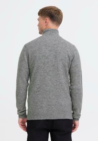 Harmaa neule pystykauluksella, joka on valmistettu pehmeästä teksturoidusta materiaalista. Varustettu pitkillä hihoilla ja istuvuudeltaan kapealla designilla, ulottuu lantioon saakka.