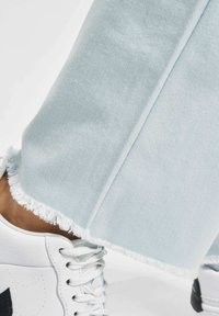 Lyseblå denimbukser med frynset nederkant, kombinert med hvite joggesko med en svart logo. Nærbilde som fremhever tekstur og detaljer.