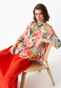 Multikleurige blouse met geometrische patronen, gemaakt van lichtgewicht stof. In combinatie met een vloeiende rode rok. Model zit op een stoel.