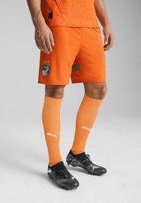 Pantalones cortos de fútbol naranja con una textura grabada, que presentan un parche con un logo y tres estrellas. Combinados con calcetas naranjas a juego y botas negras.