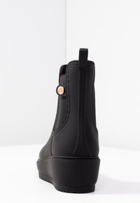 Bottine noire avec un extérieur en caoutchouc lisse, des panneaux latéraux élastiques et un talon compensé caché. Accentuée d'un logo de marque métallique rond.