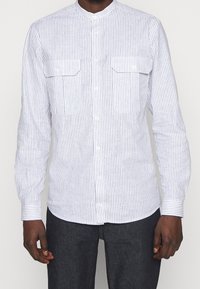 Camicia bianca a maniche lunghe con strisce verticali blu, abbottonata, con colletto alla mandarina e due tasche sul petto. Il tessuto sembra leggero e lucido.