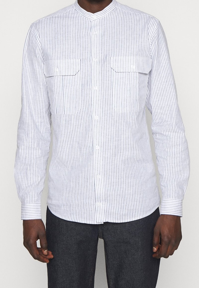 Camicia bianca a maniche lunghe con strisce verticali blu, abbottonata, con colletto alla mandarina e due tasche sul petto. Il tessuto sembra leggero e lucido.