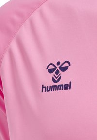 Primer plano de tela rosa con un logo negro estilizado de abeja y el nombre de la marca "hummel" impreso.