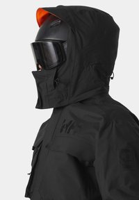 Chaqueta negra impermeable con capucha, presenta líneas angulares, bolsillos delanteros y un parche con el logo. Incluye unas gafas negras y un casco.