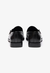 Zapatos de cuero negro con una textura suave, con un tacón bajo y sutilezas en los detalles de costura en la parte trasera. Sin herrajes visibles.