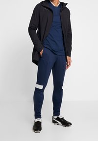 Marineblauwe sportjack over een blauwe shirt, gecombineerd met marineblauwe broek met witte accenten en zwarte voetbalschoenen, allemaal ontworpen voor prestaties.