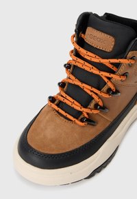 Chaussures de randonnée pour hommes Geox, en daim marron, avec des accents en caoutchouc noir et des lacets orange. Tige texturée avec un mesh respirant. Semelle épaisse.