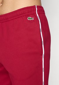 Red cotton shorts featuring an elastic waistband, side pockets, and white striped accents with "LACOSTE" text. Small embroidered logo present.