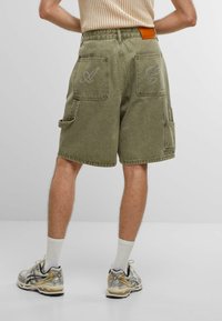 Pantaloni cargo colore verde oliva con lettere ricamate sulle tasche posteriori, caratterizzati da una vestibilità comoda e tasche laterali per utensili. Indossati con sneakers beige.