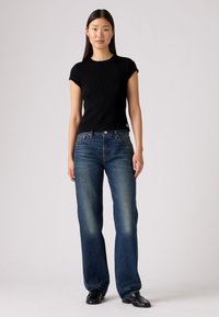 Haut côtelé noir à manches courtes associé à un jean droit bleu foncé. Le denim présente un léger délavage ; des chaussures noires élégantes complètent la tenue.