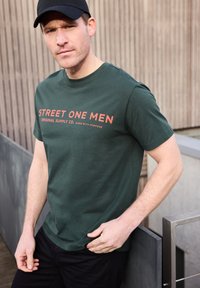 Ciemnozielona bawełniana koszulka z napisem „STREET ONE MEN” i „ORIGINAL SUPPLY CO.” w mocnym czerwonym kolorze, zestawiona z czarnymi spodniami i czarną czapką.
