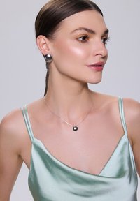 Okrągłe srebrne kolczyki i czarny naszyjnik z zawieszką noszone nad satynową miętowo-zieloną bluzką. Dominują gładka faktura i odbijające się powierzchnie.