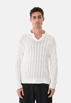 Uomo che indossa un maglione bianco lavorato a maglia con collo a V e pantaloni neri, in piedi contro uno sfondo chiaro e uniforme.