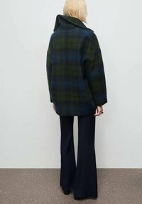 Cappotto a quadri di lana in verde scuro e blu navy, con un ampio colletto, spalle scese e una vestibilità ampia, abbinato a pantaloni scuri a zampa.