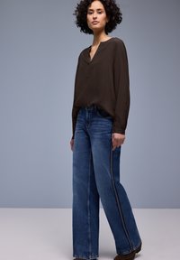 Haut marron à manches longues avec un col en V, associé à un jean bleu à jambes larges avec une bande latérale contrastante, sur un fond neutre.