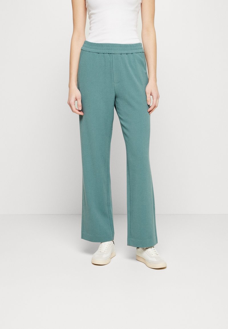 Personne portant un pantalon large turquoise, un débardeur blanc et des baskets blanches, debout devant un fond clair uni.