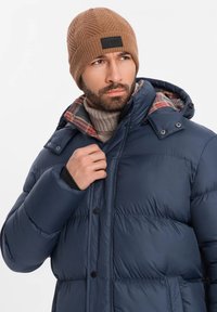 Giacca puffer blu navy con fodera a quadri, dotata di cappuccio e bottoni a pressione. Berretto a maglia marrone con motivo testurizzato e patch con logo.