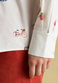 White shirt with colorful embroidered details including a bicycle and fruits, featuring a buttoned cuff and a textured red fabric underneath.
