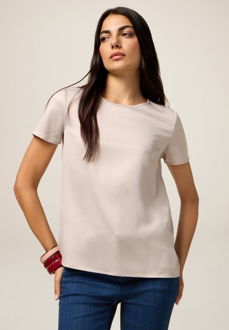 Top corto beige chiaro a maniche corte con una texture liscia, scollatura rotonda e vestibilità rilassata, abbinato a jeans blu scuro e braccialetti multicolori.