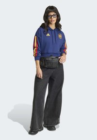 Gekürzter blauer Hoodie mit roten und gelben Streifen, Logo auf der Brust, kombiniert mit schwarzen Denim-Jeans mit weitem Bein und einer schwarzen Bauchtasche.