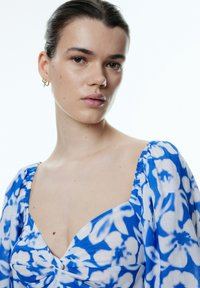 Blaue Blumenshirtbluse mit Puffärmeln, strukturiertem, leichtem Stoff, tiefem V-Ausschnitt und gerafften Akzenten an den Schultern.