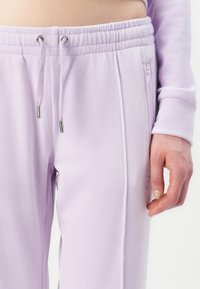 Šviesiai violetinės kelnės, pagamintos iš minkštos medžiagos, su elastingu juosmeniu su raišteliais ir užtrauktuku užsegama šonine kišene.