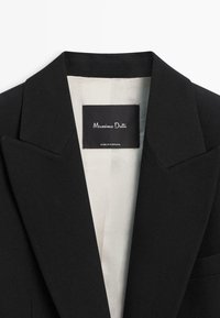Czarna marynarka z ostrymi klapami, wykonana z teksturowanej tkaniny. Wnętrze podszewki w jasnym beżu; metka z napisem "Massimo Dutti, Made in Portugal."