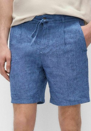 Blå chambray-shorts med forplissering og snøre i taljen, båret med en lys beige skjorte, hænder hviler ved siderne og i lommen.