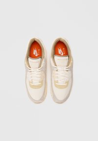 Pár béžových a krémových tenisiek Nike Air s bielymi šnúrkami a jasne oranžovými stielkami z pohľadu zhora na bielom pozadí.